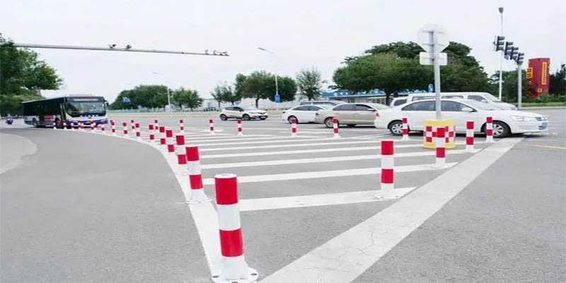White FRP marker posts with reflective bands along a curved roadway beside a public parking area.