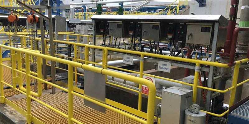 Industrial access walkway with open-grid grating, yellow guardrails, and process equipment and piping.