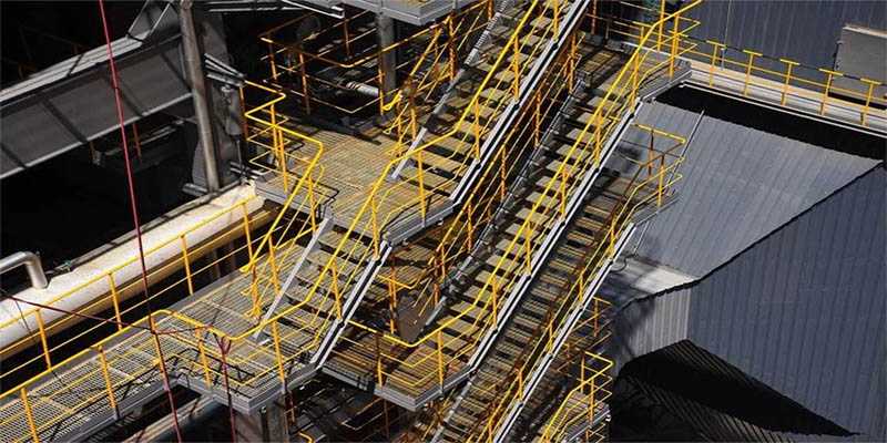 Elevated industrial walkways and access platforms with open-grid grating, stairs, and yellow safety railings around processing equipment.